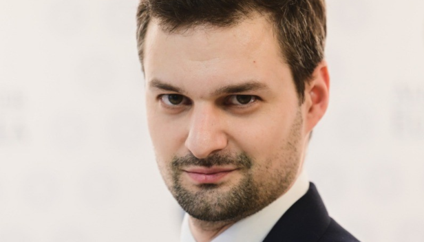 Na sliki je predstavljen moški, oblečen v profesionalno obleko, ki simbolizira strokovnost in eleganco. S temno modrim suknjičem, rdečo kravato in svetlo modro srajco, njegov videz odraža klasičen poslovni etos. Njegov samozavesten pogled in lahki nasmeh pričajo o prijetni in dostopni naravi. V ozadju je diskreten logotip z napisom "ACADEMIA", kar nakazuje, da je moški morda del akademske skupnosti ali predstavnik izobraževalne ustanove. Ta slika bi lahko bila uporabljena kot del predstavitvenega gradiva za visokošolski zavod ali kot promocija za akademski dogodek.